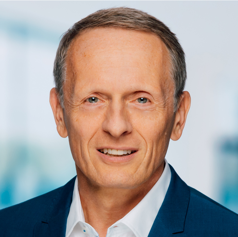 alexander-haas-portrait-hires Ein Mann mittleren Alters mit kurzem hellbraunem Haar, einem blauen Jackett und einem weißen Hemd, posiert vor einem unscharfen hellblauen Hintergrund.