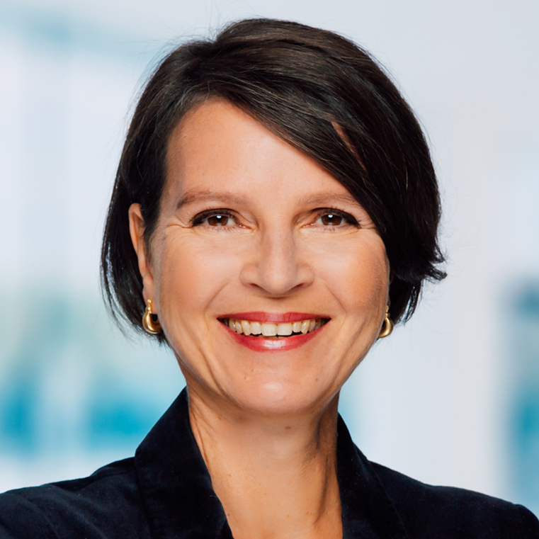 stefanie-schaefer-portrait1-hires Frau mit kurzen braunen Haaren, goldenen Creolen und einem dunklen Oberteil, lächelt vor einem unscharfen hellen Hintergrund in die Kamera.